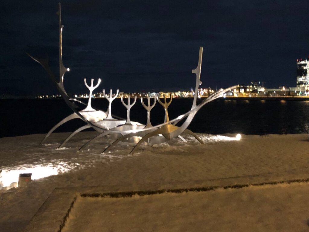 Die Skulptur Sonnenfahrt von Jón Gunnar Árnason in Reykjavik im Schnee im Dunkeln beleuchtet vor der Bucht. Im Hintergrund die Lichter der Stadt