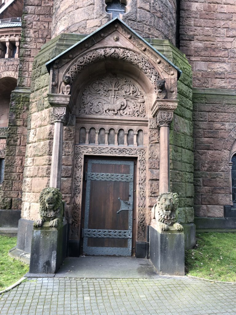 Seitenportal an der Erlöserkirche Bad Homburg. Die Tür wird von zwei liegenden Löwen umrahmt. Die Holztür ist mit eisernen Beschlägen und zwei querverlaufenden Zierbändern umfasst. Über den glatten Sandsteinsäulen gibt es ein Vordach mit einem Baldachin aus Sandstein. Darin die Abbildung des Lamm Gottes mit dem Kreuz