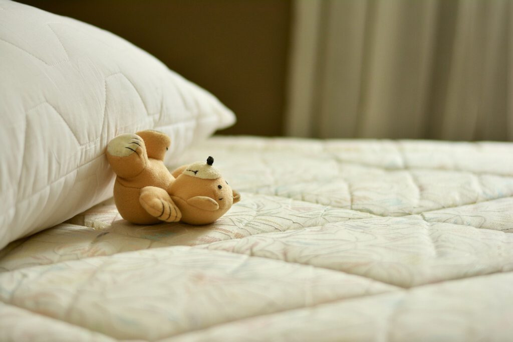 Ein hellbrauner Stoffbär liegt auf dem Rücken auf einem weiß bezogenen Bett mit den Beinen am Kopfkissen abgestützt
