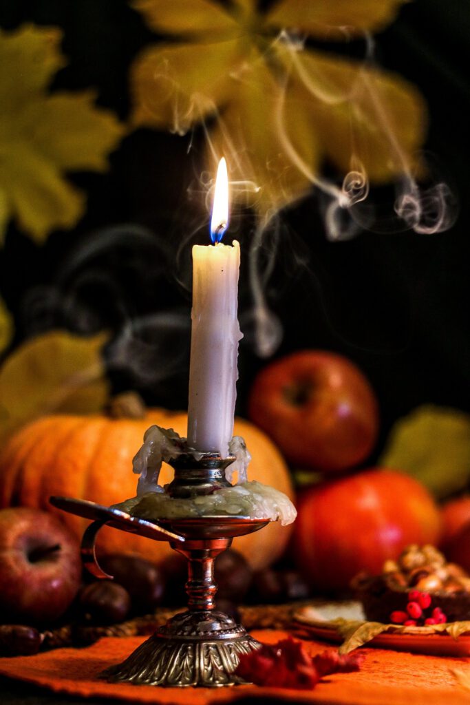 Der Rauch einer brennenden Kerze zieht ivor dem Hintergrund eines in Rot- und Orangetönen gedeckten Stilleben aus Äpfeln, einem Kürbis und gelb-grünen Blättern vor dem schwarzen Nachthimmel