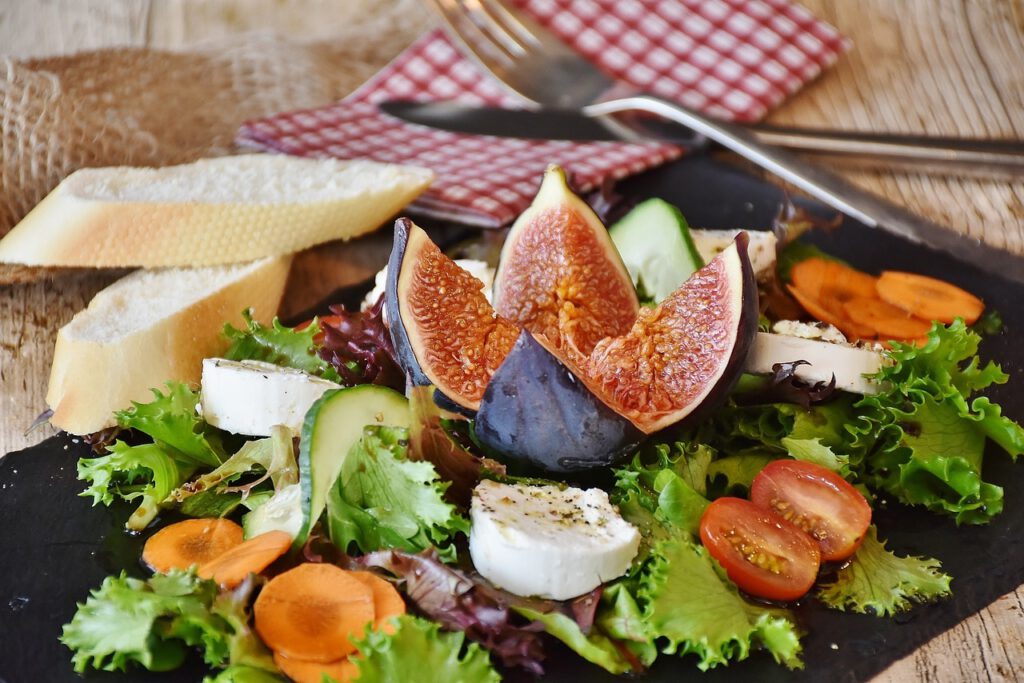 Auf einem schwarzen Schieferbrett ist ein Salat mit unterschiedlich farbigen Salatblättern, Cherry-Tomate, Gurken- und Karottenscheiben angerichtet. In der Mitte thront eine aufgeschnittene, frische Feige, um die mehrere, mit Olivenöl beträufelte, runde Scheiben Frischkäse drapiert sind. Im Hintergrund auf dem hellbraunen Holztisch befinden sich 2 Scheiben Baguette, eine rot-weiß-karierte Serviette und ein über Kreuz gelegtes Besteck.